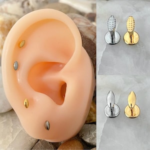 May include: Close-up of a pair of gold and silver ear studs with a textured oval design. The studs are shown on a model ear.