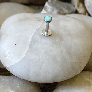 May include: A silver body piercing with a small, light blue opal gemstone.