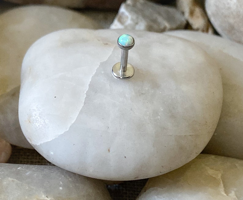 May include: A silver body piercing with a small, light blue opal gemstone.