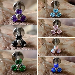 May include: A selection of six flat-back stud earrings in a titanium finish. Each earring showcases a cluster of three gemstones in diverse colours: purple, blue, pink, black, opal, green, and blue. The gemstones are arranged in a triangular formation.