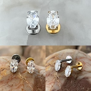May include: Oval-cut clear crystal stud earrings. One pair has a silver base, and the other has a gold base. The earrings are shown in multiple views, including close-ups, against a neutral background and a textured surface.