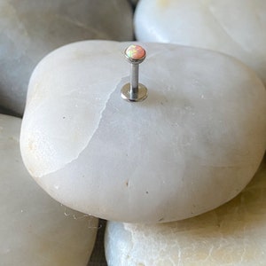 May include: A small, silver barbell with a pink opal gemstone. The barbell is on a white, smooth surface.