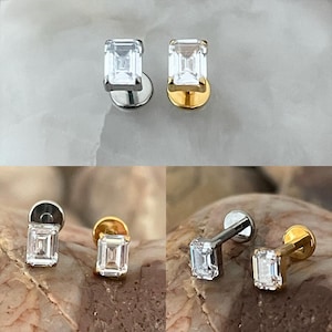 May include: Two pairs of stud earrings. One pair has a silver post and setting with a rectangular, clear gemstone. The other pair features a gold post and setting with a similar clear gemstone. The earrings are displayed on a neutral background.