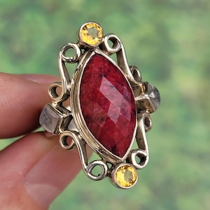 May include: A silver ring with a large red gemstone surrounded by smaller yellow gemstones. The ring has a delicate, ornate design.