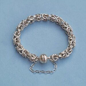 K&ouml;nnte beinhalten: Ein silbernes Kettenarmband mit einem Magnetverschluss. Das Armband besteht aus einem geflochtenen Kettenmuster.