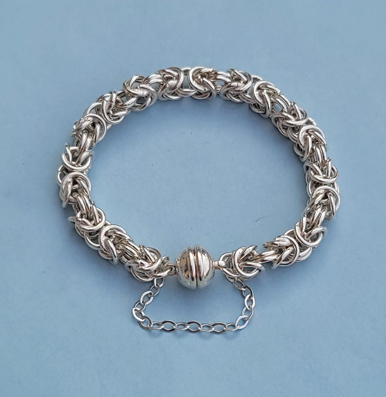 K&ouml;nnte beinhalten: Ein silbernes Kettenarmband mit einem Magnetverschluss. Das Armband besteht aus einem geflochtenen Kettenmuster.