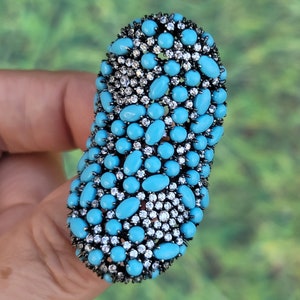 May include: A large oval-shaped ring with a cluster of turquoise and white stones set in silver. The ring is designed with a unique, textured pattern.