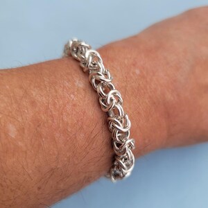 K&ouml;nnte beinhalten: Ein silbernes Kettenarmband mit einem einzigartigen, miteinander verwobenen Design. Das Armband wird am Handgelenk einer Person gezeigt.