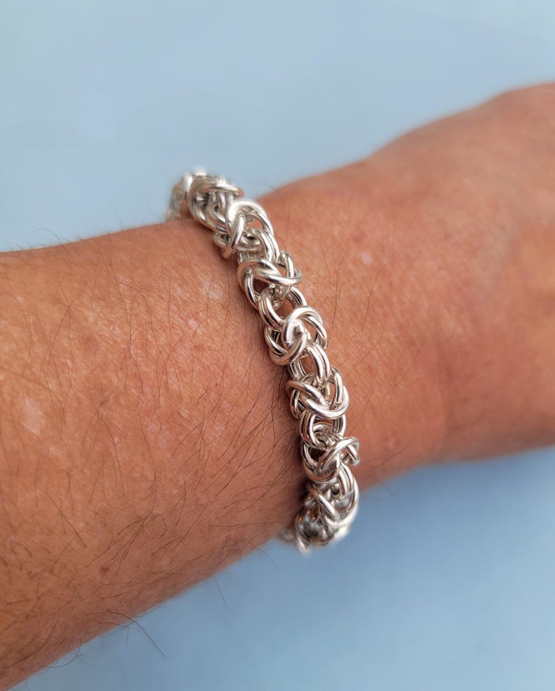K&ouml;nnte beinhalten: Ein silbernes Kettenarmband mit einem einzigartigen, miteinander verwobenen Design. Das Armband wird am Handgelenk einer Person gezeigt.