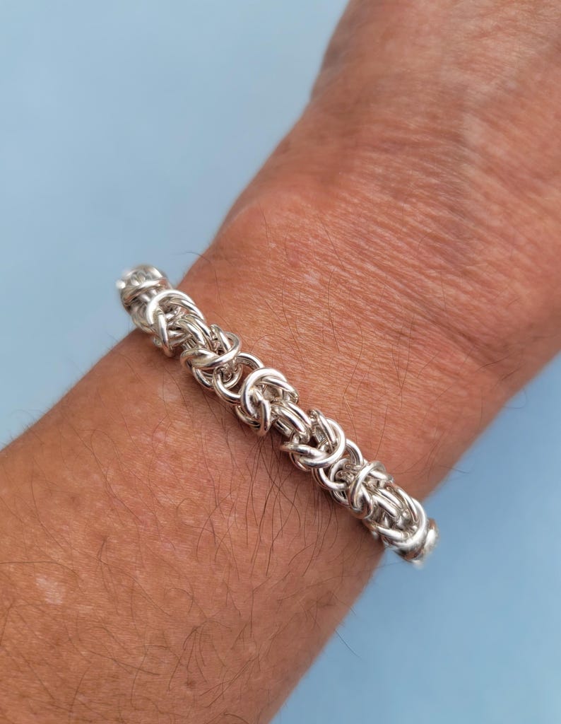 K&ouml;nnte beinhalten: Ein silbernes Kettenarmband mit geflochtenem Design. Das Armband befindet sich an einem Handgelenk.