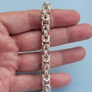 K&ouml;nnte beinhalten: Ein silbernes Kettenarmband mit geflochtenem Design. Das Armband wird auf einer Hand gezeigt.