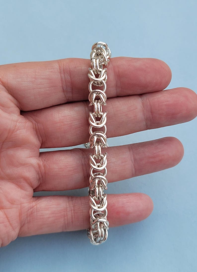 K&ouml;nnte beinhalten: Ein silbernes Kettenarmband mit geflochtenem Design. Das Armband wird auf einer Hand gezeigt.