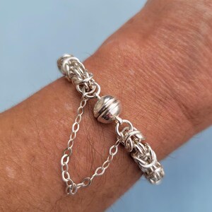 K&ouml;nnte beinhalten: Ein silbernes Armband mit einem magnetischen Verschluss. Das Armband besteht aus einem geflochtenen Kettenmuster.