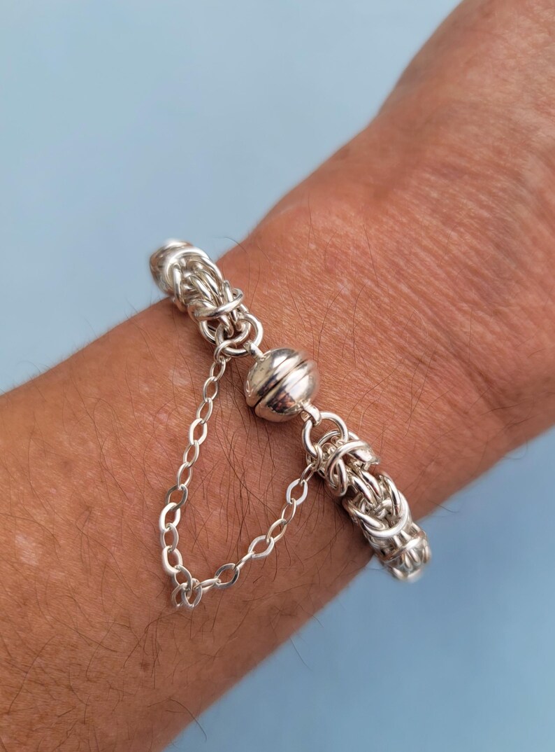 K&ouml;nnte beinhalten: Ein silbernes Armband mit einem magnetischen Verschluss. Das Armband besteht aus einem geflochtenen Kettenmuster.