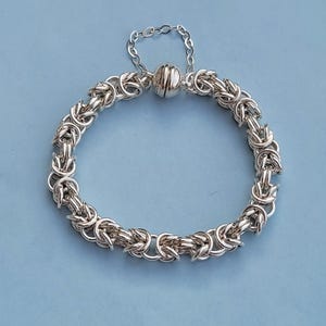 K&ouml;nnte beinhalten: Ein silbernes Kettenarmband mit einem Magnetverschluss. Das Armband besteht aus einem geflochtenen Kettenmuster.