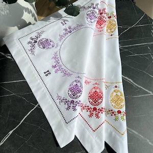 Può includere: Tre tovaglioli di lino bianco con ricami di uova di Pasqua in viola, rosso e oro. Ogni tovagliolo presenta una diversa combinazione di colori e accenti floreali.