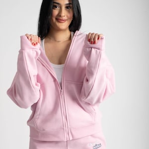 Könnte beinhalten: Ein rosa Kapuzenpullover mit Reißverschluss und eine passende rosa Jogginghose. Die Jogginghose hat die Worte "Old Nine" auf dem linken Bein gestickt.