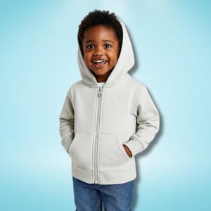 May include: A pale grey zip-up hoodie with a hood and front pockets. The hoodie features a small embroidered design on the chest. The child is wearing blue jeans and smiling.