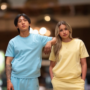 Puede incluir: Dos personas vestidas con camisetas y pantalones de chándal de colores pastel a juego. Una persona lleva un conjunto azul claro, la otra un conjunto amarillo. Cada camisa tiene un pequeño diseño de nube en el pecho. El fondo está borroso con tonos cálidos.