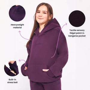 May include: A deep purple heavyweight hoodie featuring a kangaroo pocket with a tactile sensory fidget patch, long sleeves with built-in stress balls in the cuffs, and a hood. The garment includes embroidered text details on the chest and pants.