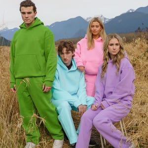 May include: Four people wearing matching hooded sweatshirts in different colors: green, pink, light blue, and purple. They are standing in a field with mountains in the background.
