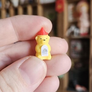 Può includere: Una bottiglia di miele gialla in miniatura a forma di orso con un tappo rosso. La bottiglia ha un'etichetta bianca con la scritta "HONEY". L'orso ha gli occhi neri ed è tenuto in mano.