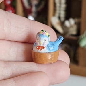 May include: A small, blue bird ornament with a floral crown and a red bow, sitting in a tiny brown basket. The bird has black eyes and is intricately painted. This miniature item is likely for a doll's house or a collector's display.