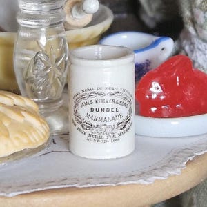 Pode incluir: Um pote miniatura de cerâmica branca de marmelada James Keiller & Sons Dundee com texto impresso em preto, posicionado ao lado de um pequeno objeto vermelho translúcido num prato branco, um pastel e um vaso de vidro transparente.