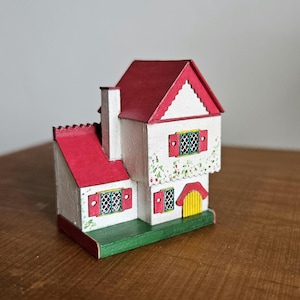 May include: A small, white, two-story dollhouse with a red roof and green base. The house has red shutters and a yellow door. The walls are decorated with a floral pattern.