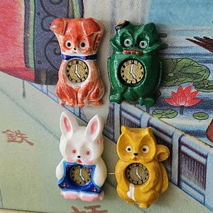 May include: Four whimsical animal-shaped clocks: a red dog, a green frog, a white rabbit, and a yellow owl. Each clock has a circular face with black hands and a gold-colored frame. The clocks are set against a colorful background.