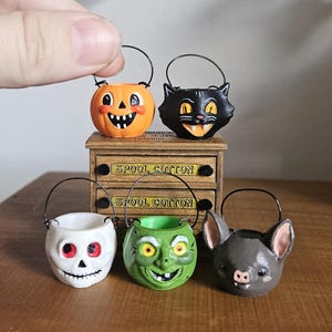 Może przedstawiać: Pięć miniaturowych wiaderek na Halloween z czarnymi drucianymi uchwytami, przedstawiających dynię, czarnego kota, białą czaszkę, zieloną czarownicę i szarego nietoperza. Stoją na małej drewnianej szafce z dwiema szufladami z napisem SPOOL COTTON.