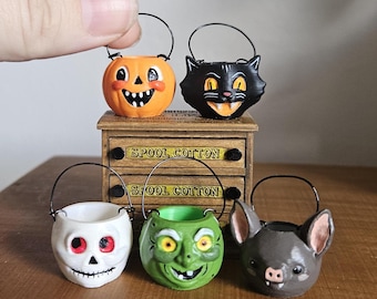 May include: Five miniature Halloween buckets with black wire handles, featuring a pumpkin, black cat, white skull, green witch, and grey bat. They are displayed on a small wooden cabinet with two drawers labeled SPOOL COTTON.
