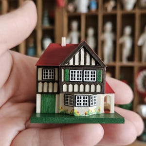 May include: A miniature house model with a red roof, brown trim, and white walls. The house features multiple windows with green shutters and a green door. The model sits on a green base with painted flowers. The house is held in a hand.