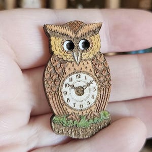 May include: A wooden owl-shaped clock with a detailed design. The clock face is visible within the owl's body, showing the numbers and hands. The owl is primarily brown with yellow and white accents, and sits on a green base.