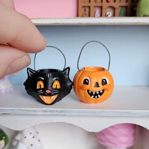 May include: Two miniature Halloween candy buckets: a black cat and an orange jack-o'-lantern. Both have smiling faces and silver wire handles. The cat bucket has orange accents, and the pumpkin bucket has black accents.