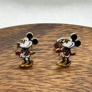 Może przedstawiać: Para złotych kolczyków z motywem Myszki Miki. Kolczyki przedstawiają kreskówkową postać Myszki Miki w czerwono-białym stroju.