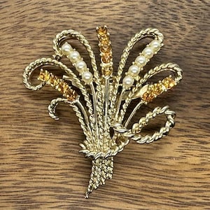 May include: A gold-toned brooch in the shape of a wheat sheaf, adorned with faux pearls and amber-colored rhinestones. The brooch has a textured, detailed design, and is set against a wooden background.