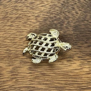 May include: A gold-colored turtle brooch with a lattice shell design. The brooch has small, sparkling accents and is set on a wooden surface. The turtle's head and flippers are detailed.
