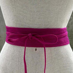 Może przedstawiać: Tkany pas w kolorze magenta zawiązany w kokardę. Pas jest owinięty wokół manekina. Paski pasa zwisają. Pas jest wykonany z teksturowanego materiału. Tło to betonowa ściana.