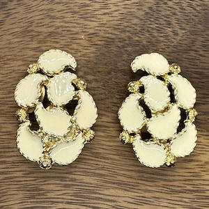 May include: A pair of cream-colored clip-on earrings with a gold-tone metal setting. Each earring features a cluster of leaf-shaped enamel pieces, accented with small gold-colored beads. The earrings are displayed on a wooden surface.