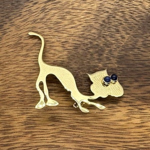 May include: A gold-toned cat brooch with blue gemstone eyes. The cat is in a playful pose with its tail raised.