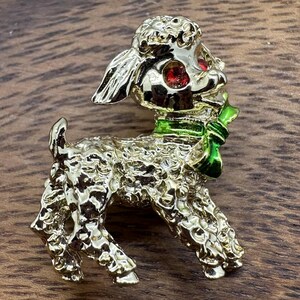 May include: A gold-toned lamb brooch with red eyes and a green scarf. The lamb has a textured, curly coat and is standing on all fours. A vintage-style accessory.