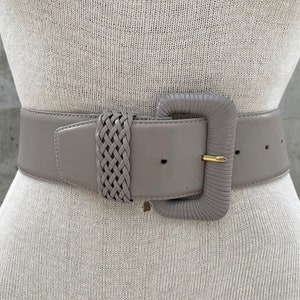 Puede incluir: Un cintur&oacute;n ancho de cuero gris con una hebilla grande y cuadrada. La hebilla tiene un dise&ntilde;o trenzado y un acabado dorado.