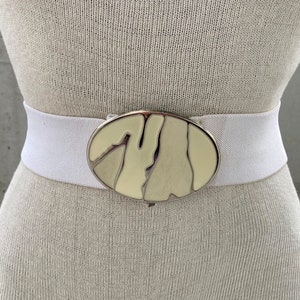 Puede incluir: Cinturón elástico blanco con una hebilla ovalada grande. La hebilla es de metal y tiene un diseño de color crema.