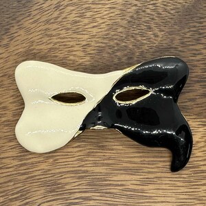 May include: A decorative mask-shaped brooch, split into cream and black sections. The brooch has gold-colored accents and eye holes. The mask is set on a wooden surface.