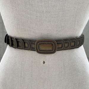 May include: A brown metal belt with a large, ornate, rectangular buckle. The buckle has a rope-like design around the edge and a smooth, flat center.