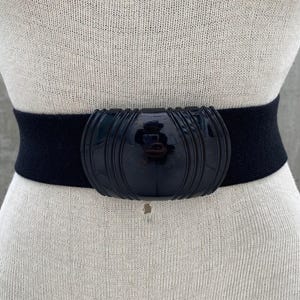 Puede incluir: Un cinturón elástico negro con una hebilla grande de plástico negro. La hebilla tiene una forma ovalada redondeada con un diseño texturizado y acanalado.