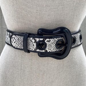 May include: A black and white snakeskin patterned belt with a large, black, glossy buckle. The buckle has a unique, curved shape.
