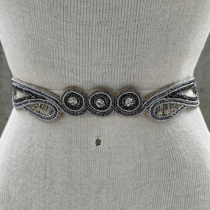 May include: A beaded belt with a cream-colored base. The belt features a central design of four circular embellishments with silver and clear beads. The sides of the belt have a decorative pattern with silver and gold beads. The belt is displayed on a mannequin.