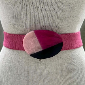 May include: A pink elastic belt with a large oval buckle. The buckle is divided into three sections: pink, black, and a marbled pink and white. The belt is displayed on a mannequin.